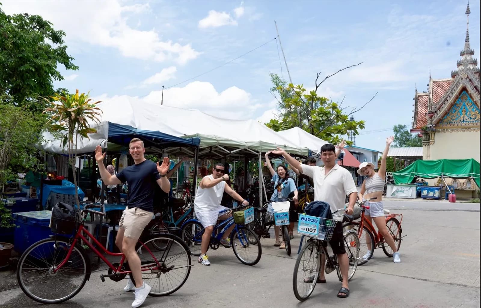 An O-World Travel guide and a small group of guests waving from their bicycles beside a local market and temple
